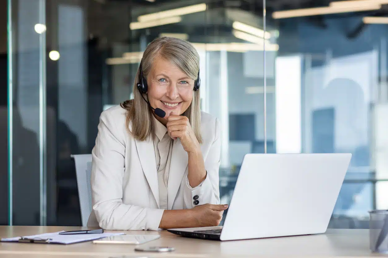 Portrait of a smiling senior gray-haired business woman sitting at a desk in the office in front of a laptop, wearing a headset and confidently looking at the camera.